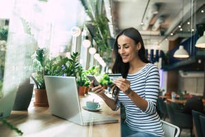 Smiling young woman using laptop and doing shopping online with help of her smartphone and card